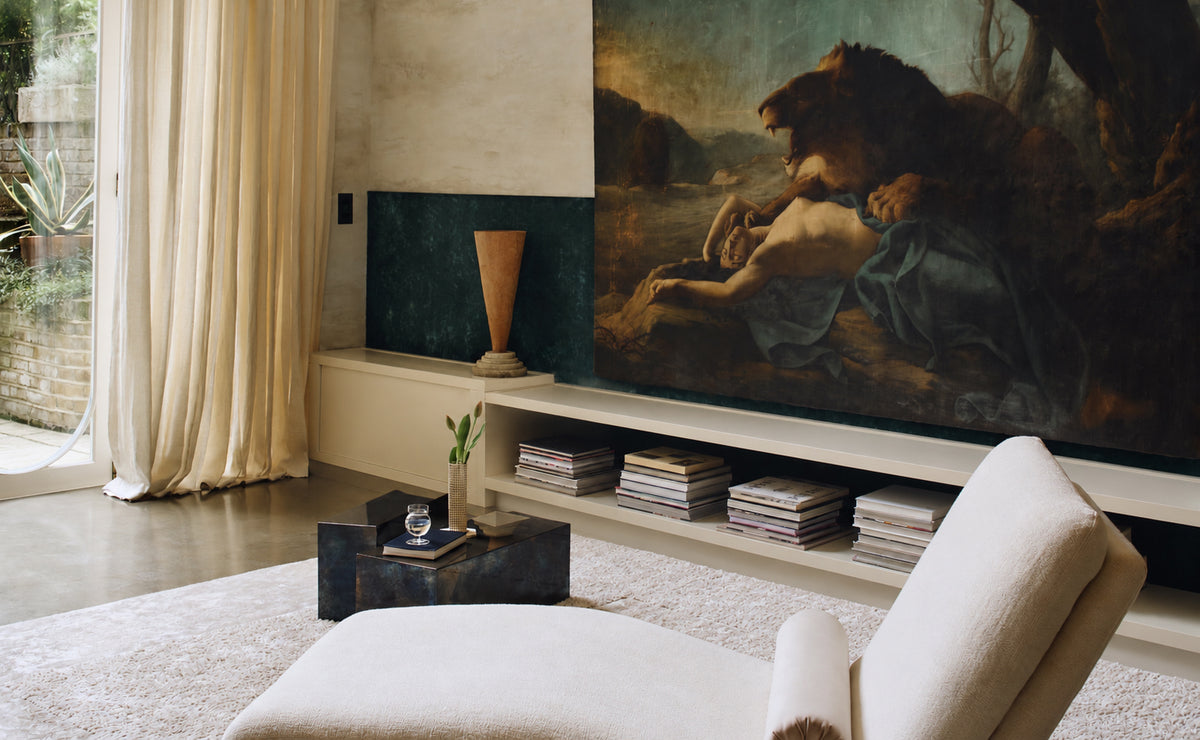 A large classical-style painting depicting a lion and a reclining figure, displayed on a wall in a modern living room setting with a beige armchair, console table, and curtains.