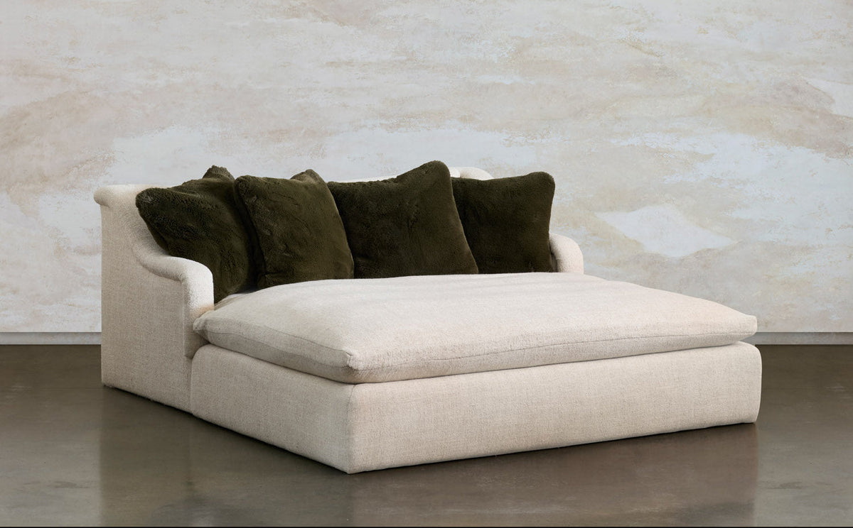 A modern low-profile daybed or chaise lounge upholstered in light beige fabric, styled with four dark olive green velvet throw pillows, set against a textured beige plaster wall and a dark concrete floor.