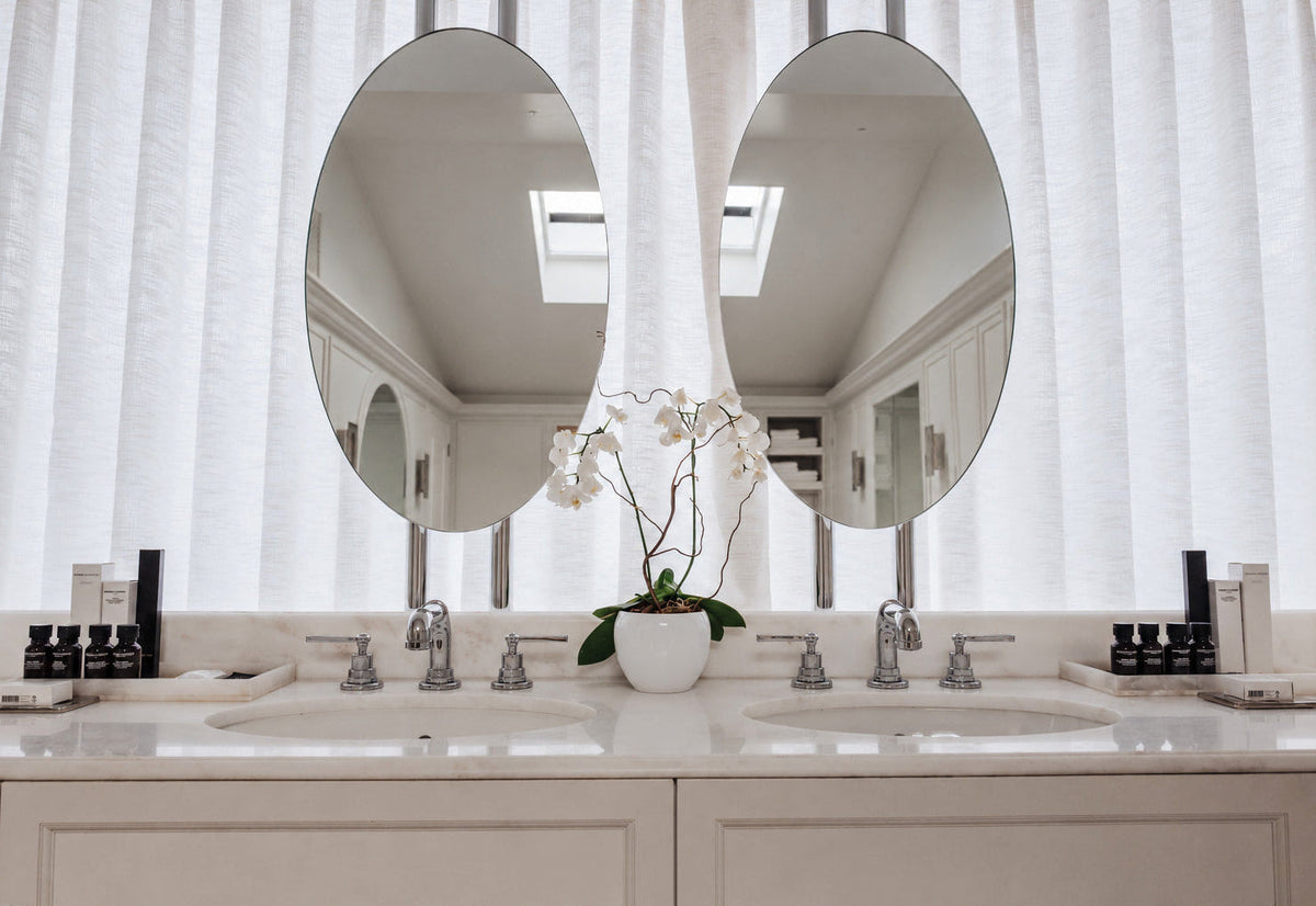 A pair of large oval wall mirrors mounted above a double bathroom vanity with white sinks and chrome faucets, set against white vertical blinds and reflected in the mirrors.
