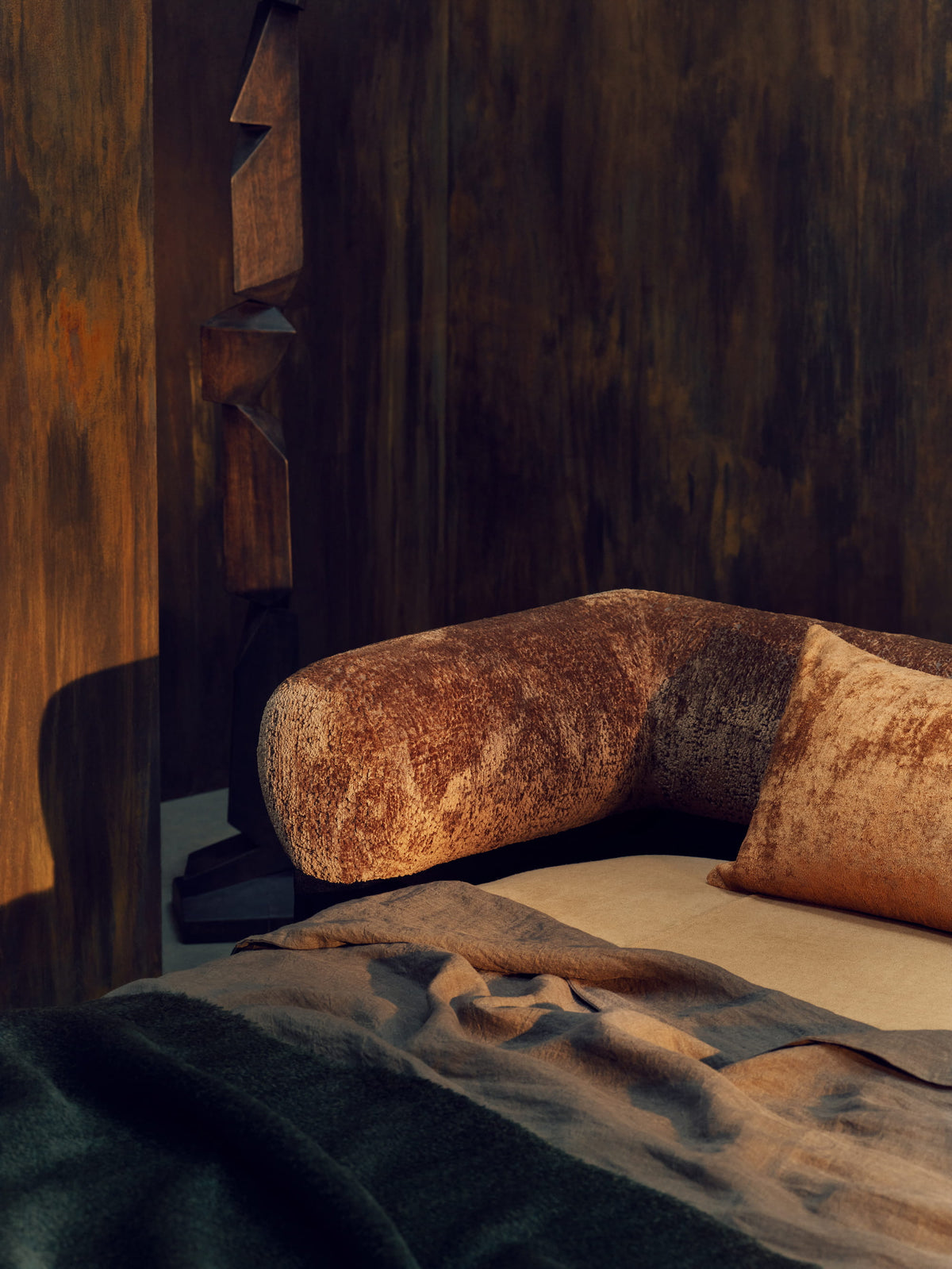 Detail of the Nest bed’s rust velvet bolster beside a carved wooden sculpture and rumpled linen bedding