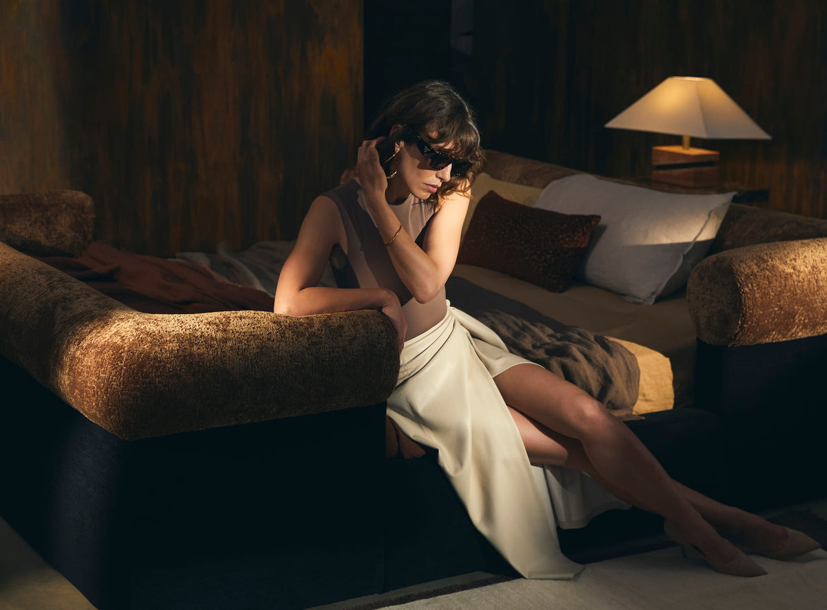 A woman in a cream dress seated on the Nest bed, with rust velvet bolsters and a lit table lamp