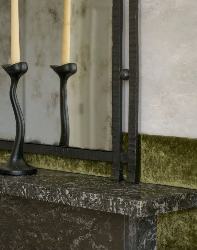 A close-up of a dark metal fireplace mantel with a vertical bar and a matching candlestick holder, set against a textured gray wall and green velvet trim.