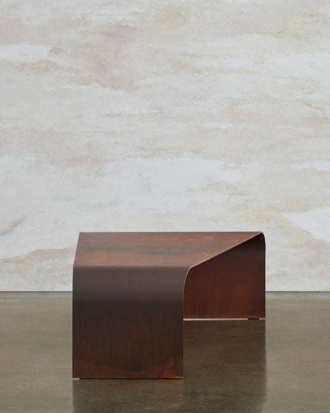 A modern sculptural side table made of rusted copper-colored metal, set against a textured beige plaster wall and a polished concrete floor.