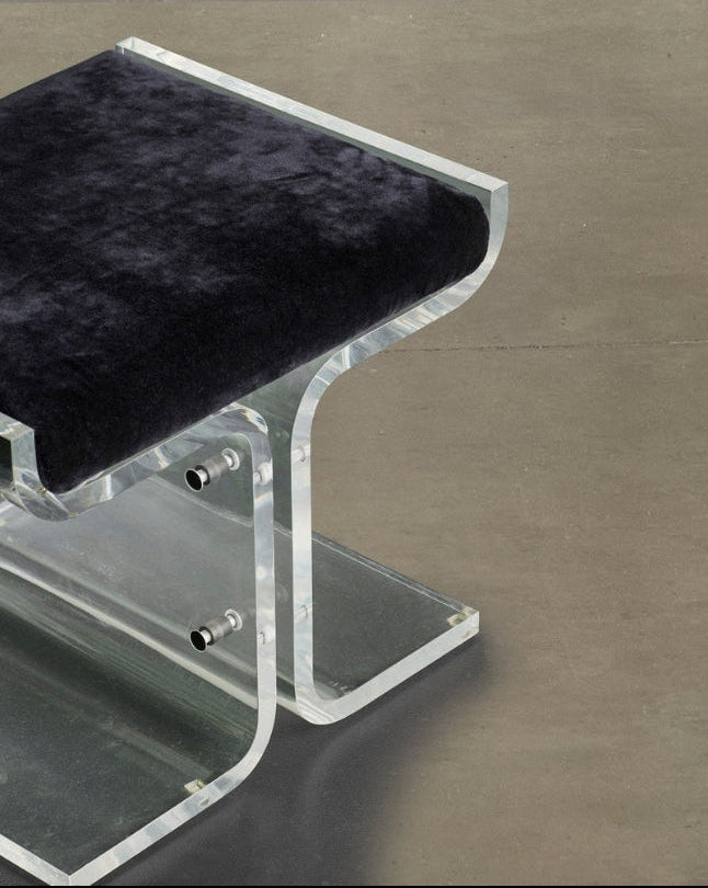 A modern acrylic stool with a curved, transparent clear-acrylic frame and a plush black velvet seat, shown on a neutral concrete floor.