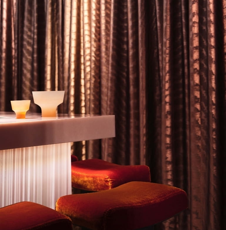 Backlit fluted bar with terracotta velvet stools and bronze drapery at CoOc Members Club