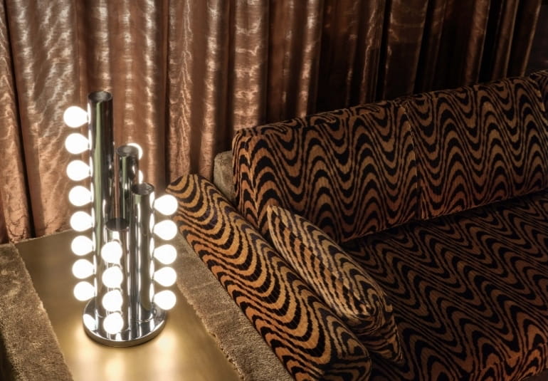 Multi-bulb chrome lamp beside a tiger-stripe sofa and copper curtains at CoOc Members Club