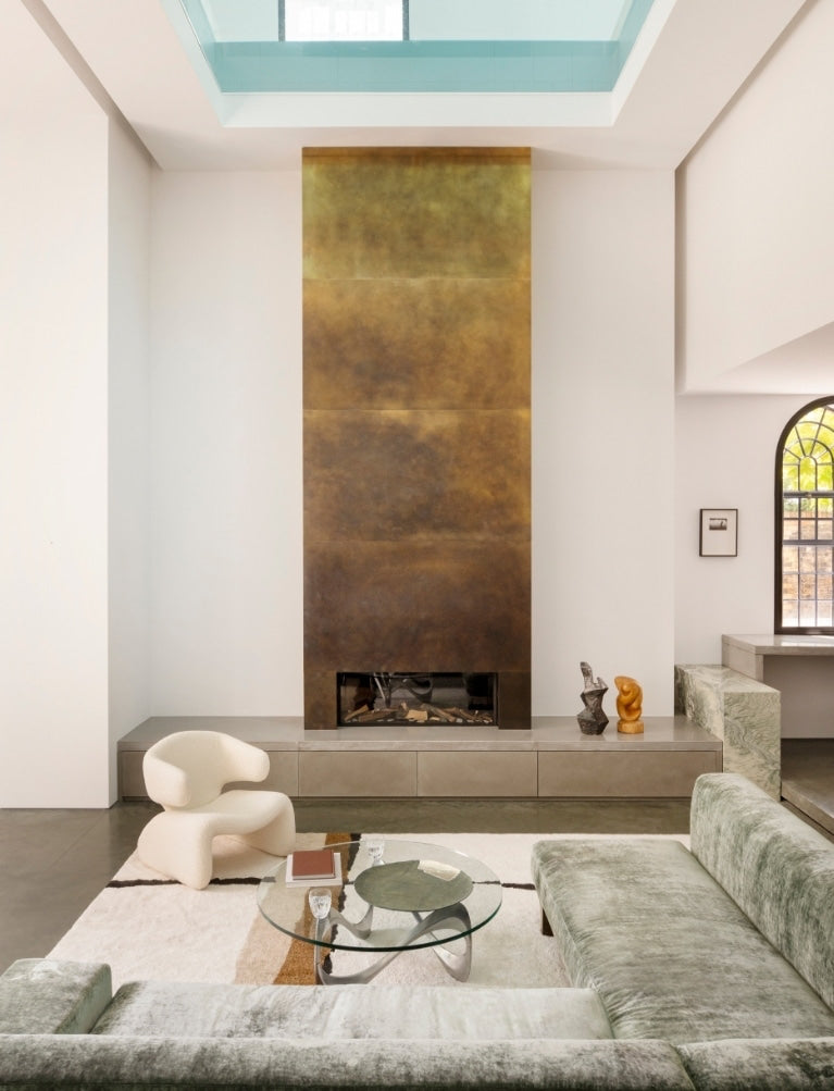 Patinated brass fireplace under a skylight with a sage velvet sofa in the North London warehouse